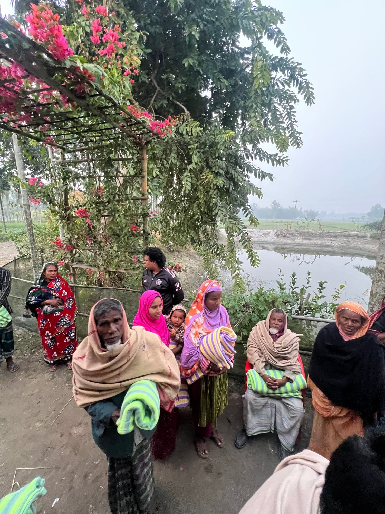 Families gathered with blankets beside farmland in Rangpur